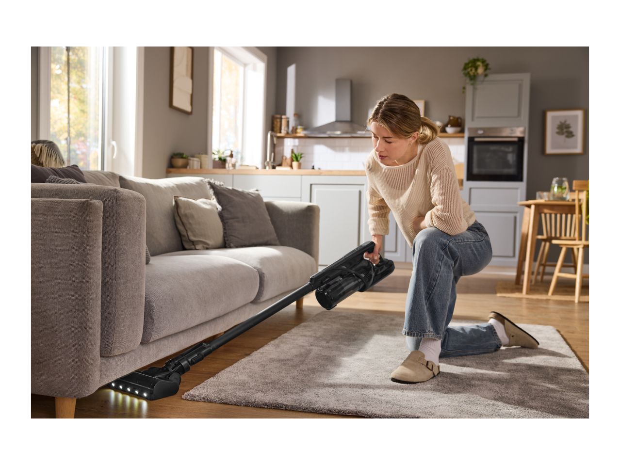 Woman vacuuming under a sofa with a cordless stick vacuum cleaner in a modern living room.