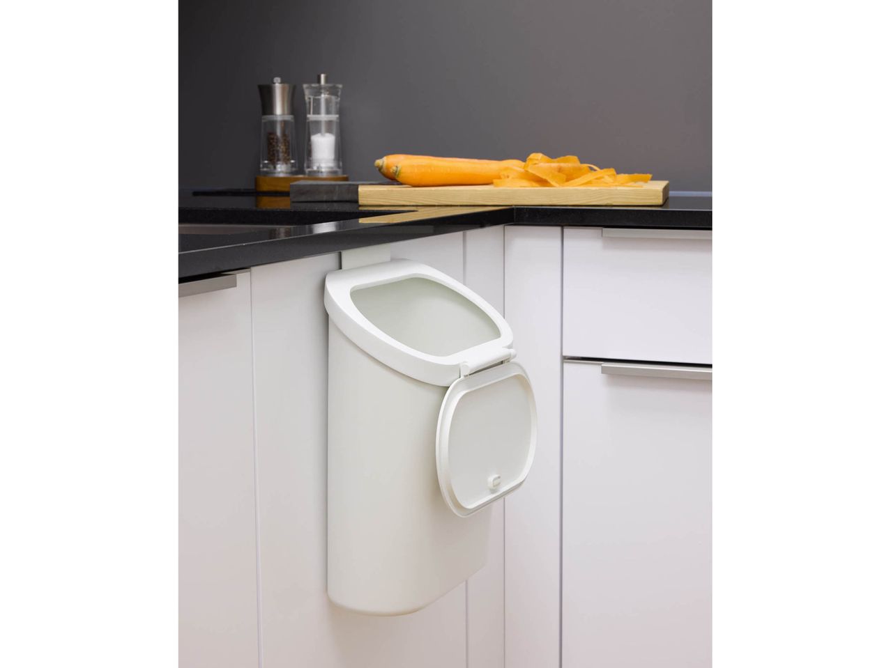 White compost bin hanging from a kitchen counter with a carrot on a cutting board.