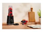 A black personal blender filled with berries next to a bowl of fresh fruit on a wooden counter.