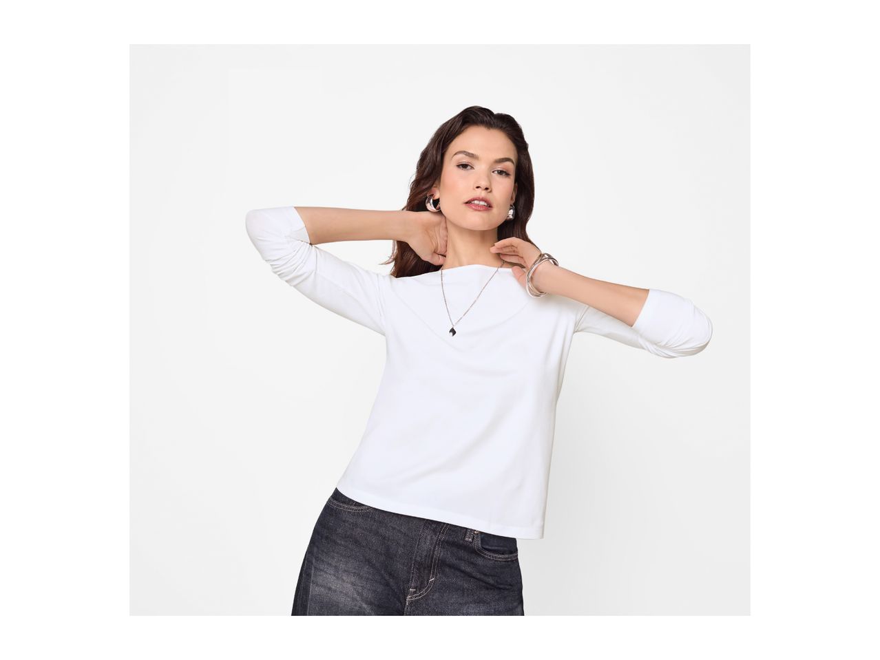 Woman in a white long-sleeved top and dark jeans, wearing silver hoop earrings, a necklace, and bracelets.