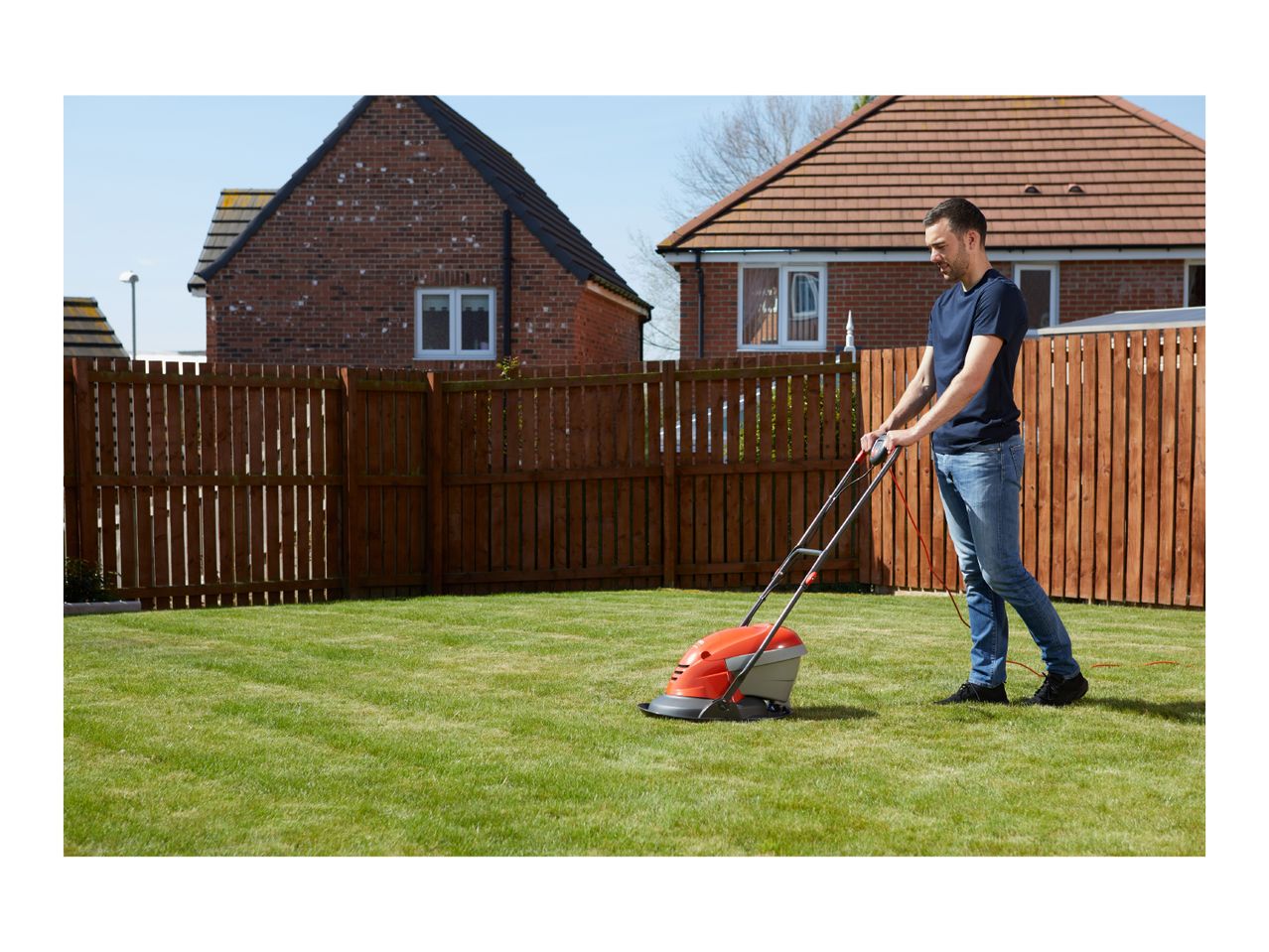 Man mowing a green lawn with a red and grey lawnmower in a garden with a wooden fence.