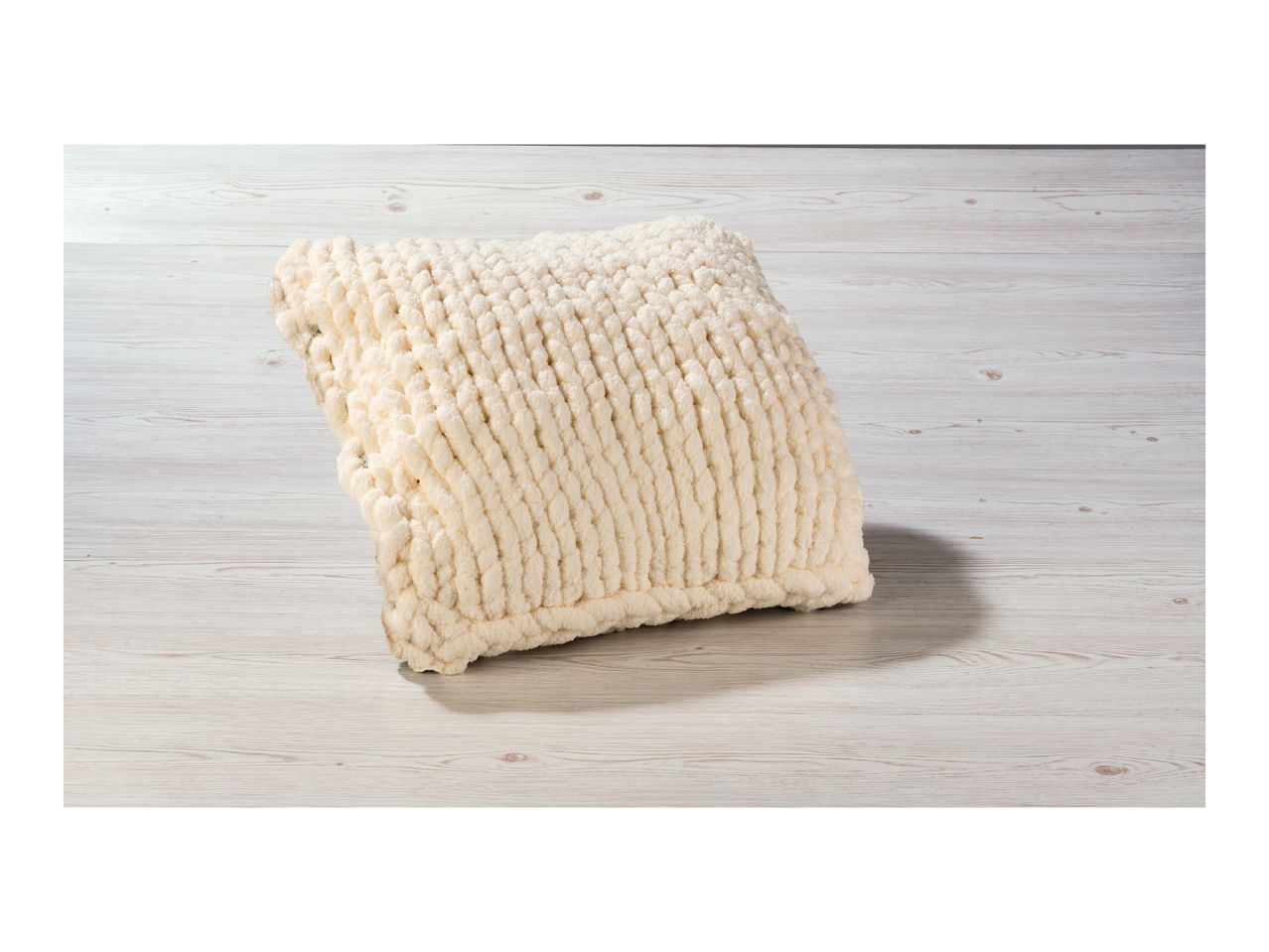 A cream-colored chunky knit blanket or throw pillow on a light wooden surface.
