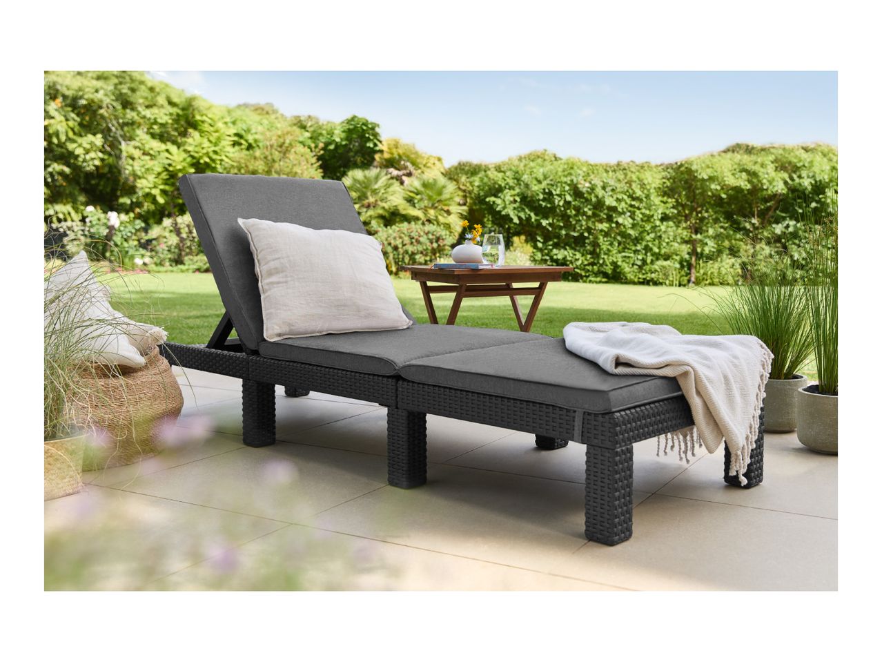 Dark grey sun lounger with cushions and a blanket on a patio, surrounded by greenery.