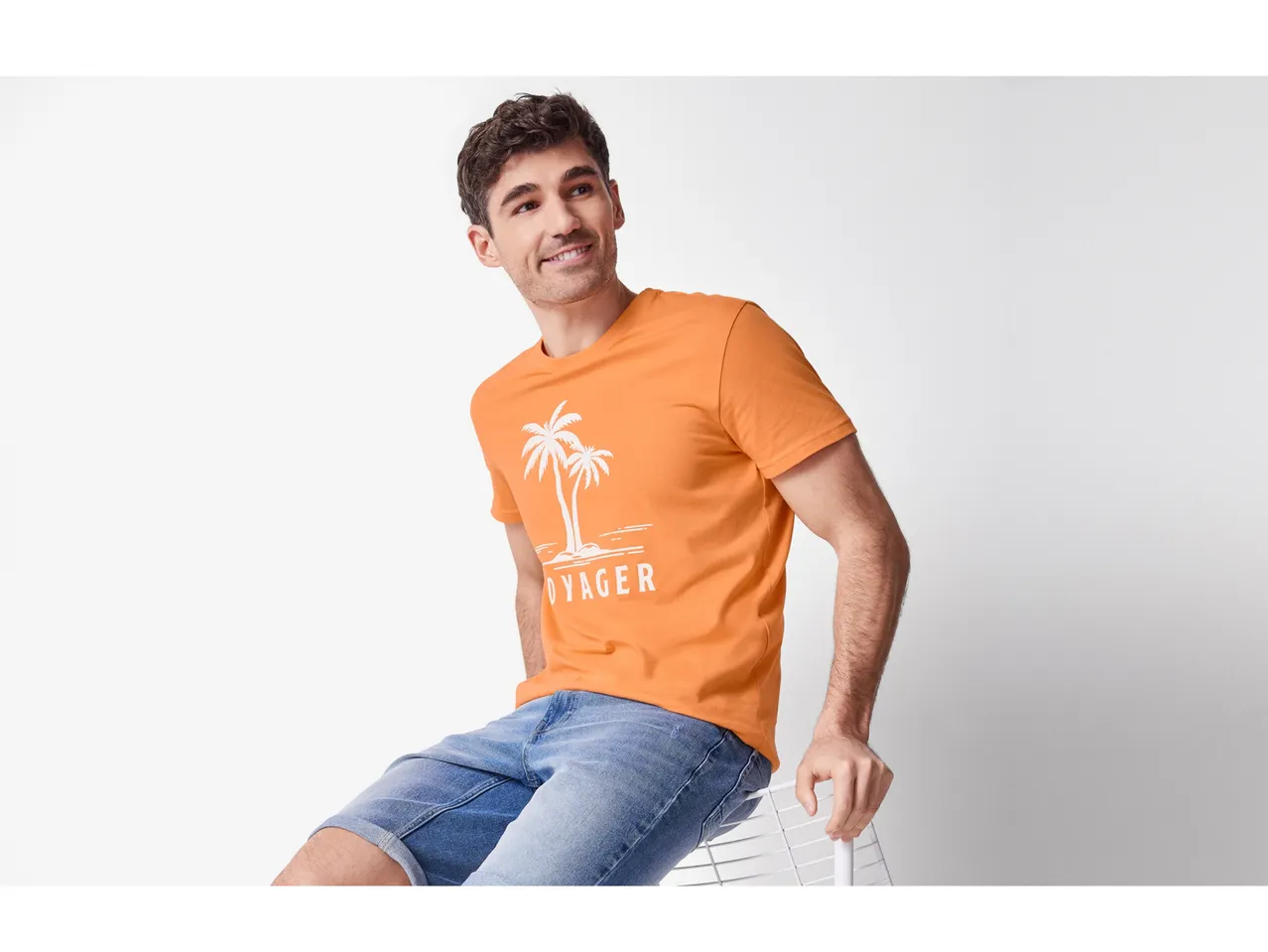 Model masculin îmbrăcat într-un tricou portocaliu cu imprimeu cu palmieri și pantaloni scurți din denim.