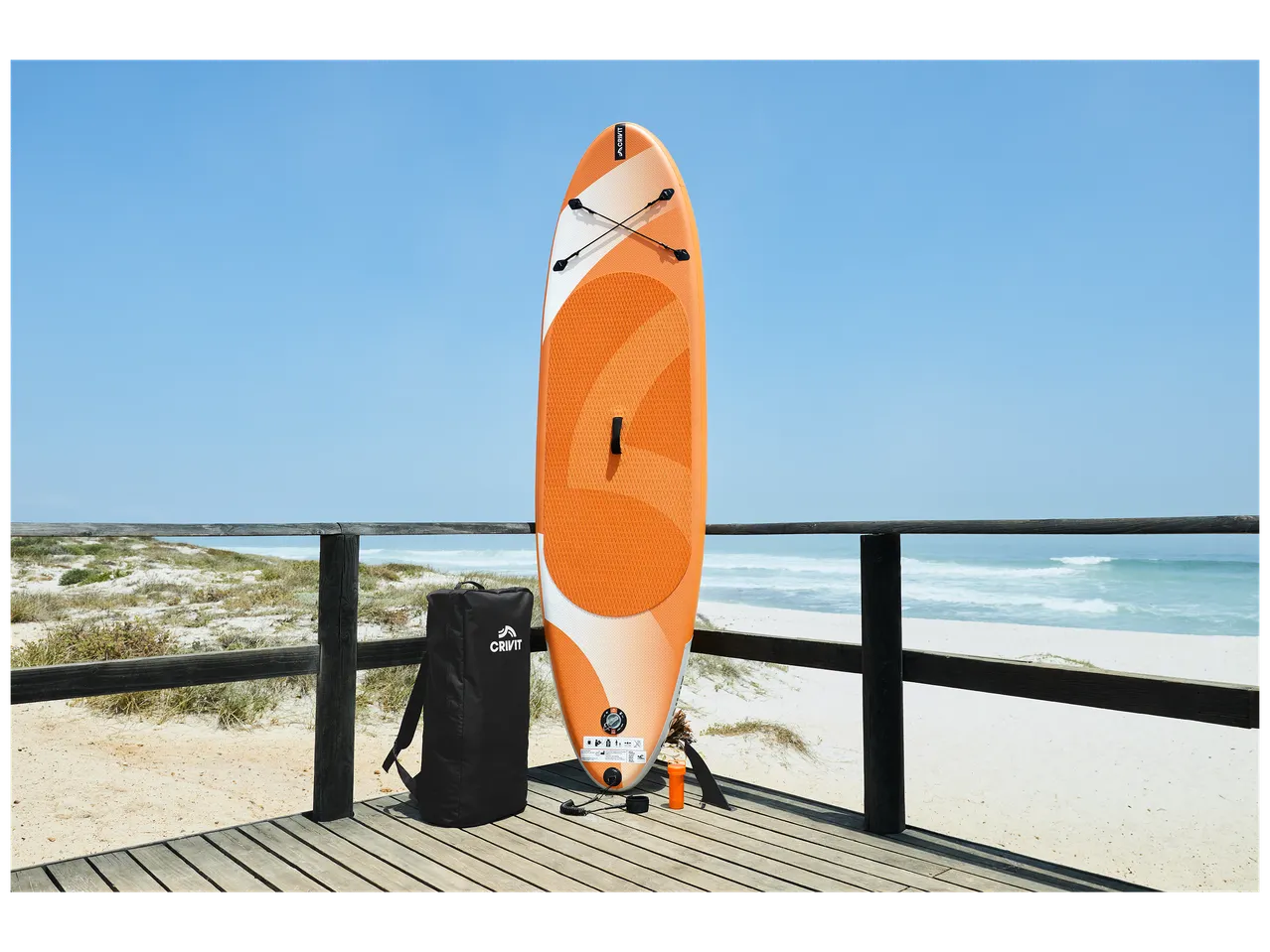 Planche de paddle orange et blanche avec un sac à dos noir sur une terrasse en bois face à la mer.