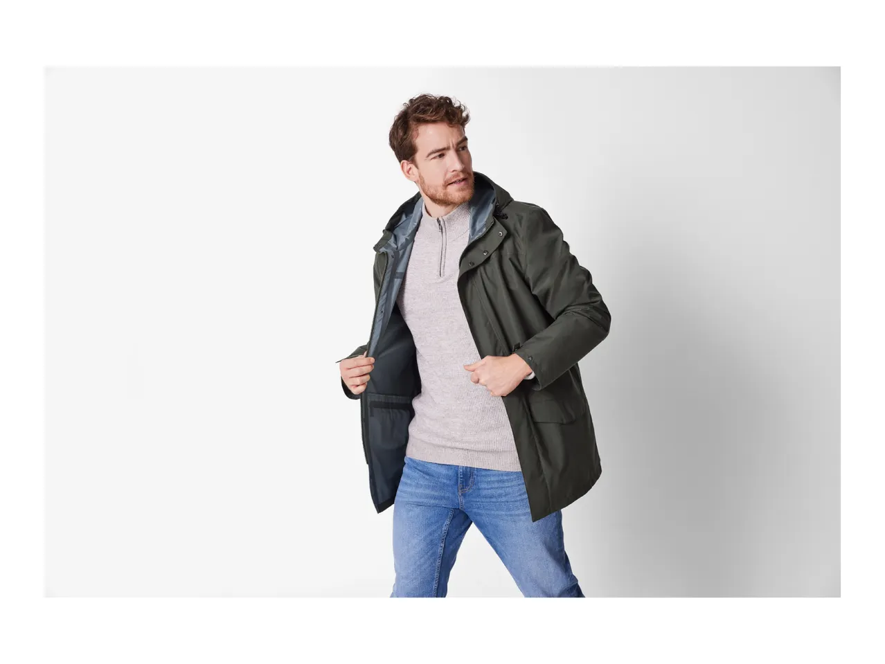 A man in a dark green jacket and blue jeans looks to the side.