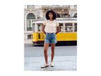Woman in a cream top and denim shorts, standing on a street with a yellow tram passing by.
