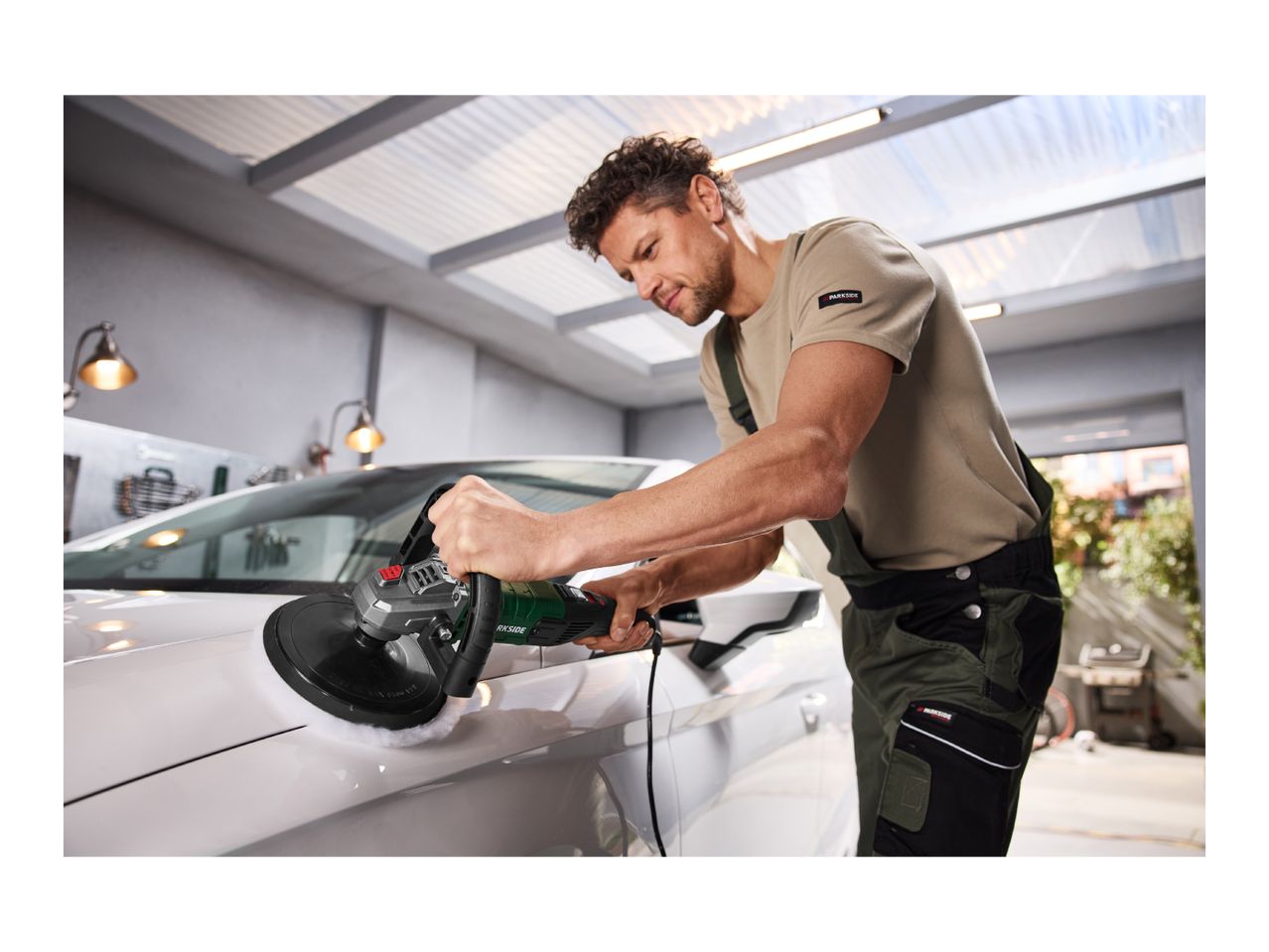 Man polishing a white car with an electric polisher in a garage.