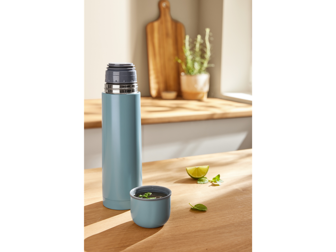 A light blue thermos and a cup with mint tea on a wooden kitchen counter.