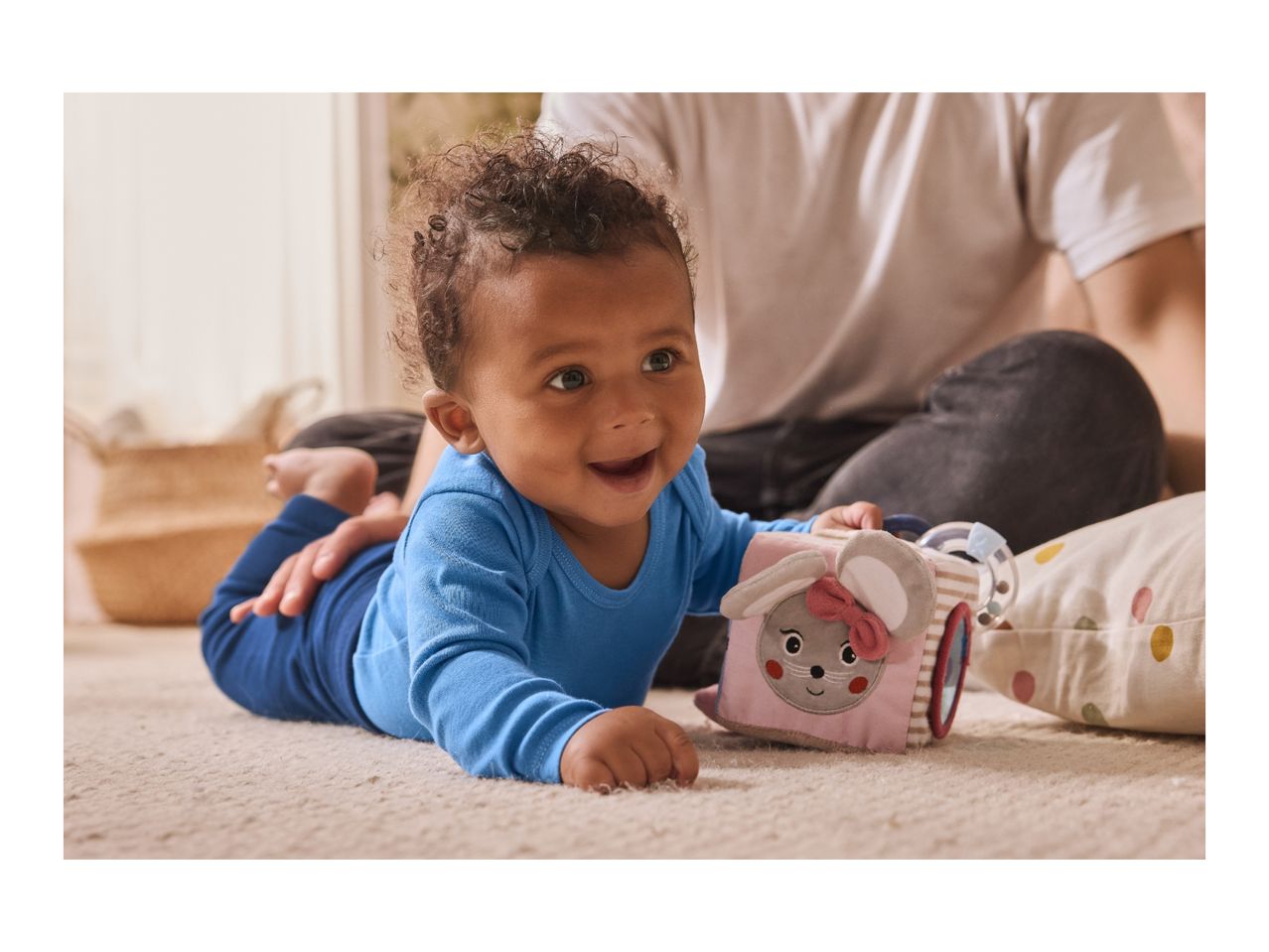 Happy baby in blue outfit playing with a soft toy cube on a carpet.