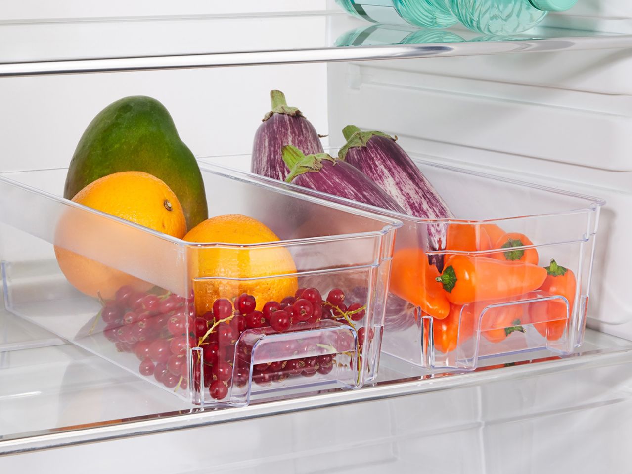Clear plastic refrigerator bins filled with fresh produce like mango, oranges, eggplants, and peppers.