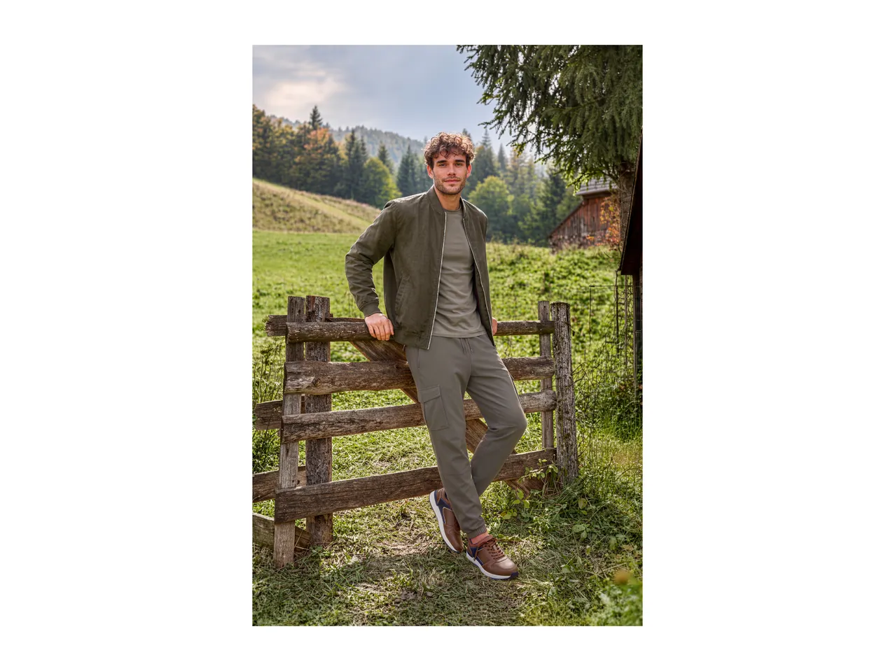 Man in olive green bomber jacket, t-shirt, and cargo pants, leaning on a wooden fence in a field.