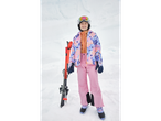 Smiling girl in a floral ski jacket, pink ski pants, and a white helmet with goggles, holding skis.