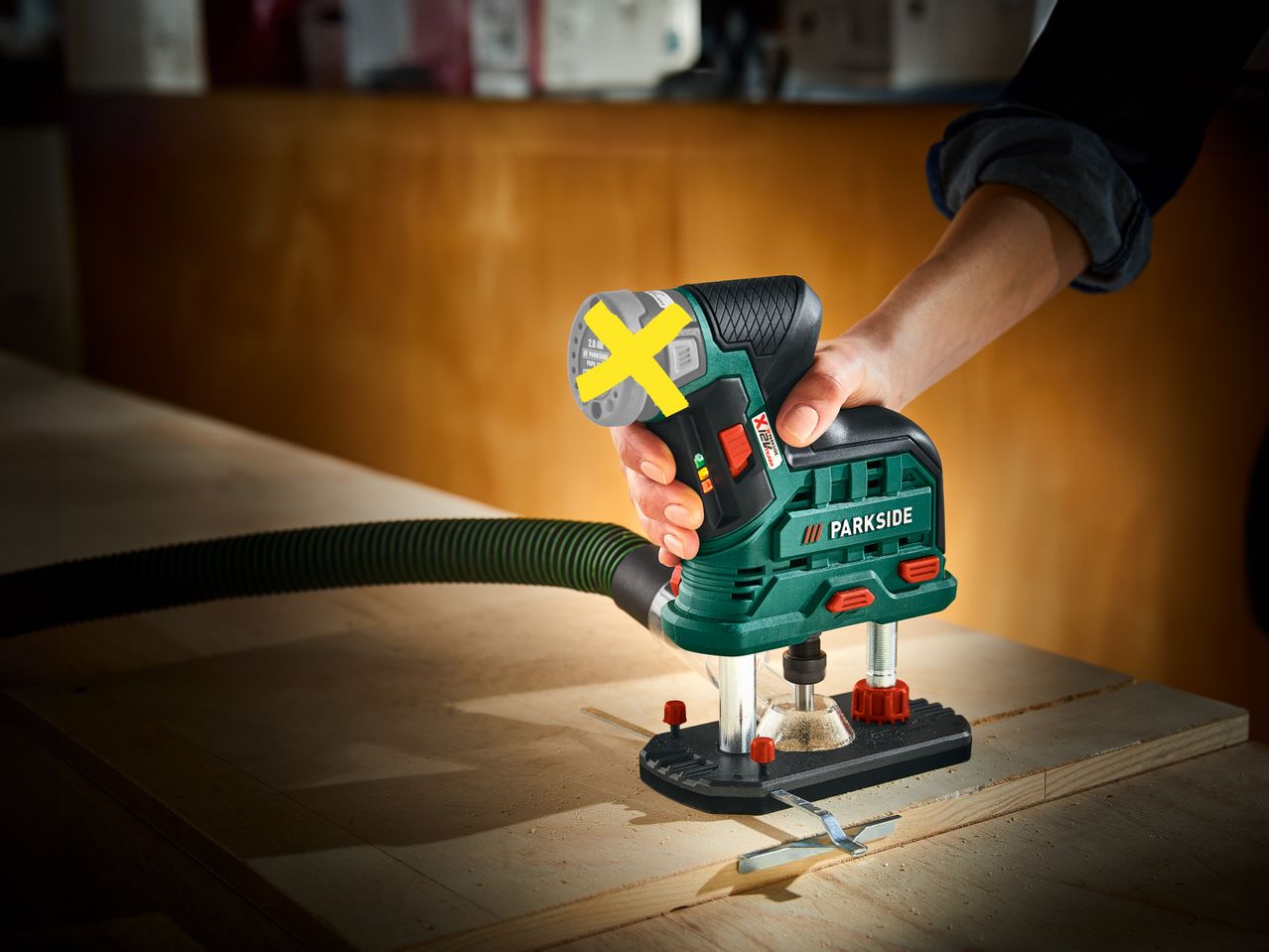 Una persona usa una Fresatrice verticale ricaricabile Parkside su un pezzo di legno, con un tubo di aspirazione collegato.