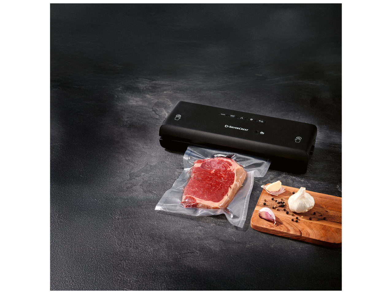 Appareil de mise sous vide avec un steak emballé, de l'ail et des grains de poivre sur une planche en bois.