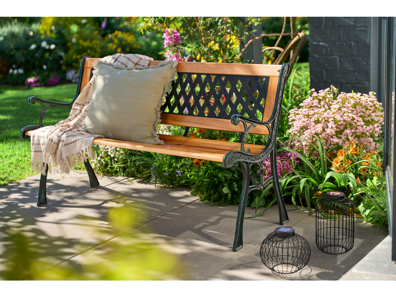 Livarno Garden Bench with a cushion and throw, surrounded by lush garden plants.