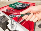 A hand uses a ratchet wrench on a metal part near a red toolbox filled with tools.