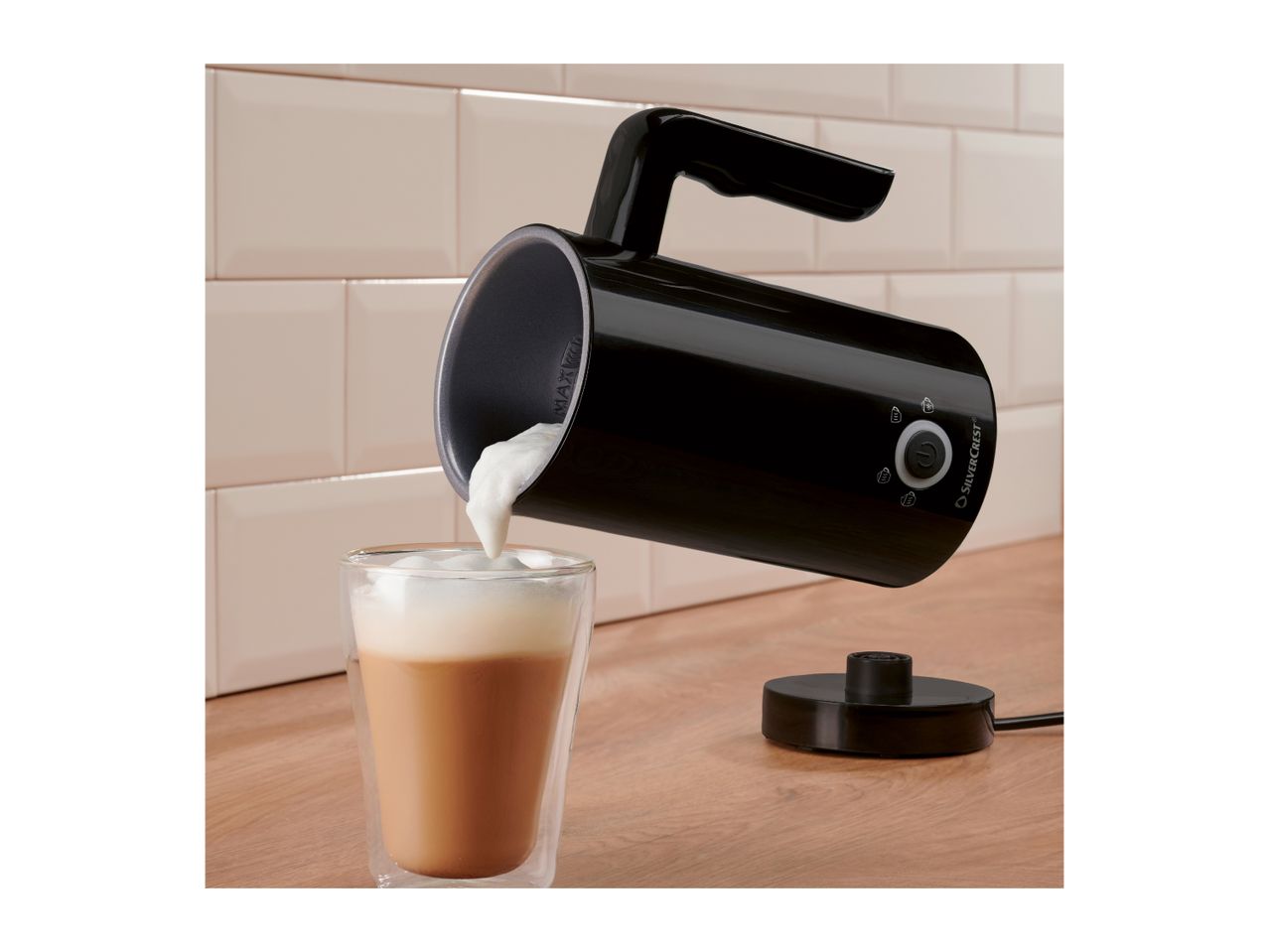 Black milk frother pouring foam into a glass of latte on a wooden counter.