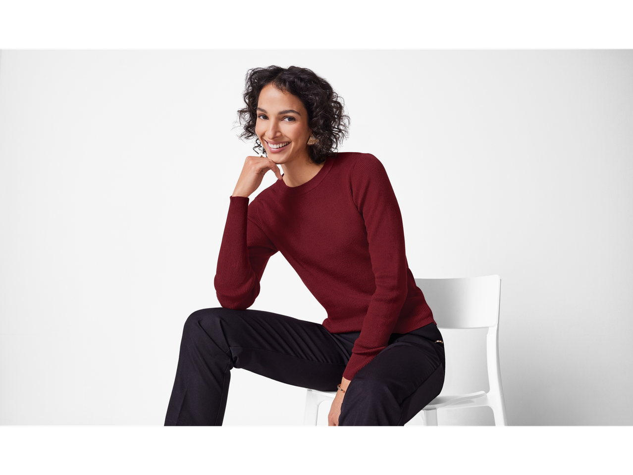 Nasmijana žena u bordo puloveru i crnim hlačama, sjedi na bijeloj stolici.