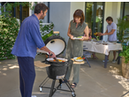 Man grilling vegetables on a ceramic barbecue, woman holding a plate of food.