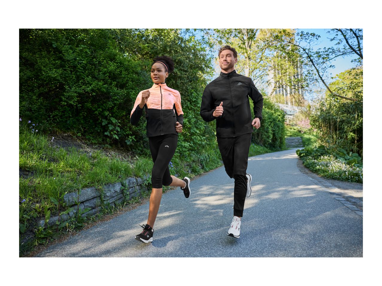 Two people jogging outdoors in athletic jackets and pants on a sunny day.