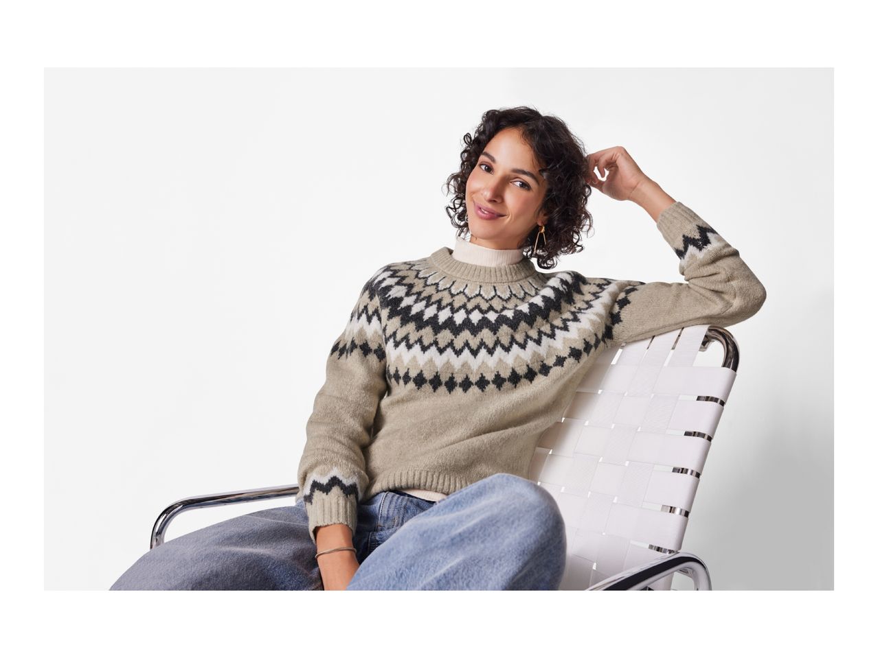 A smiling woman in a patterned knit sweater and jeans sits in a white chair.