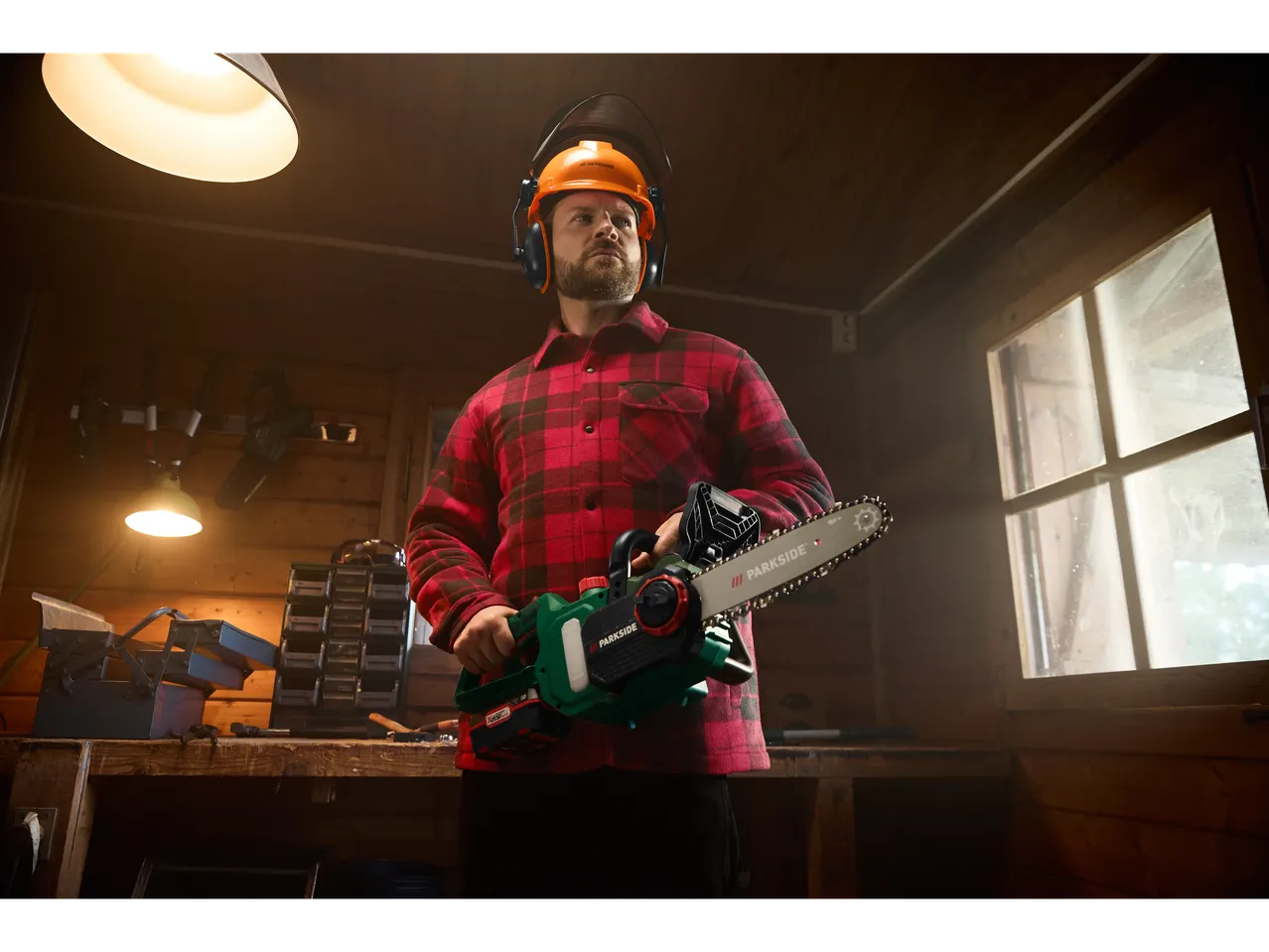Man in Parkside helmet and ear defenders holding a Parkside chainsaw in a workshop.