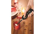 A hand holds a long lighter, igniting a candle among others on a wooden surface.