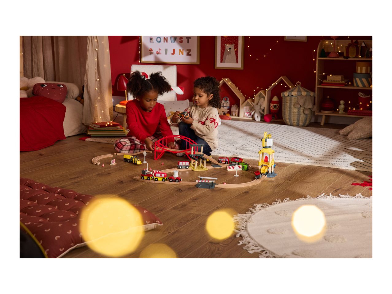 Two children playing with a wooden train set and toy vehicles in a festive room.