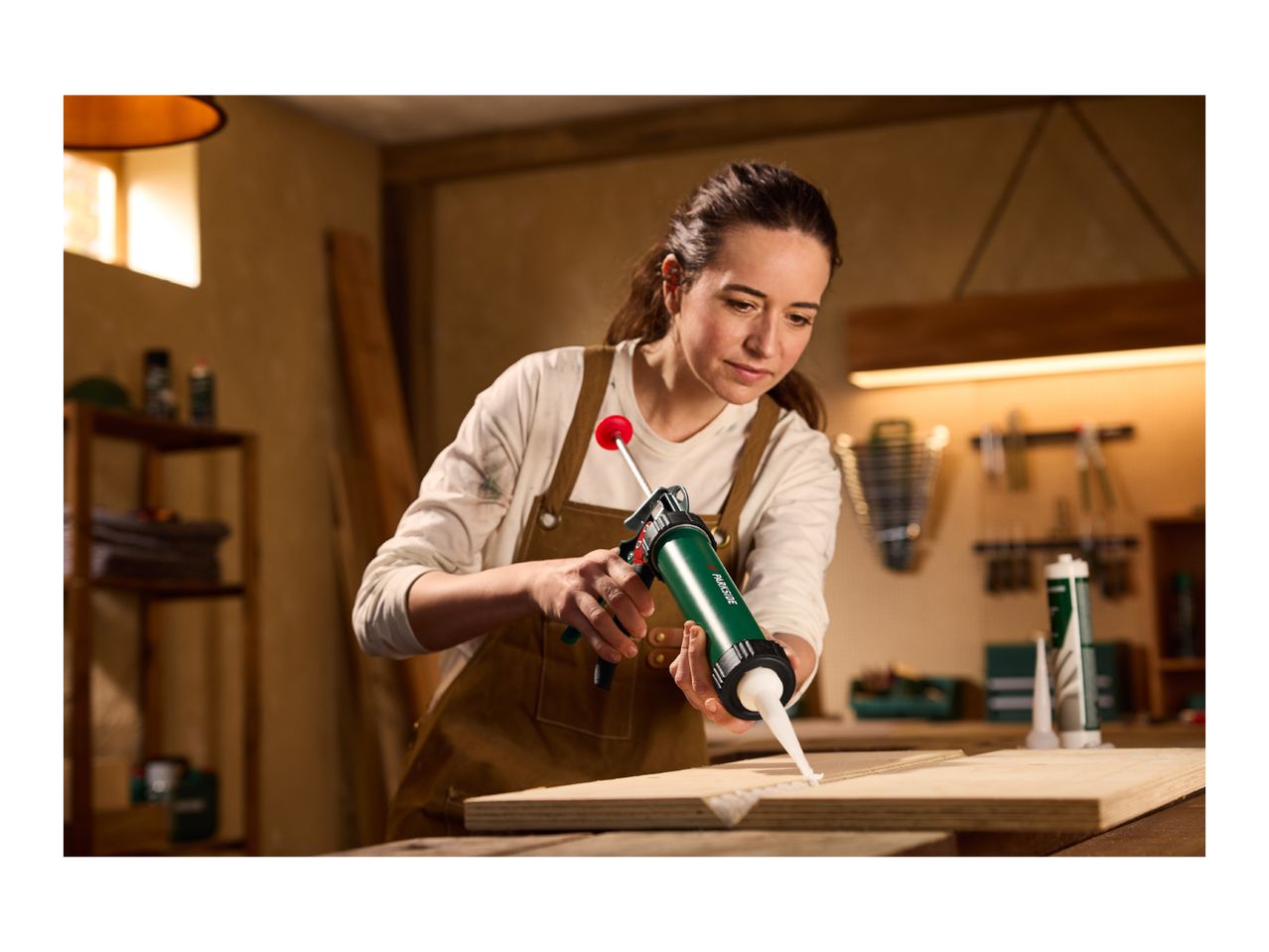 Woman applying sealant with a caulk gun to a wooden board in a workshop.