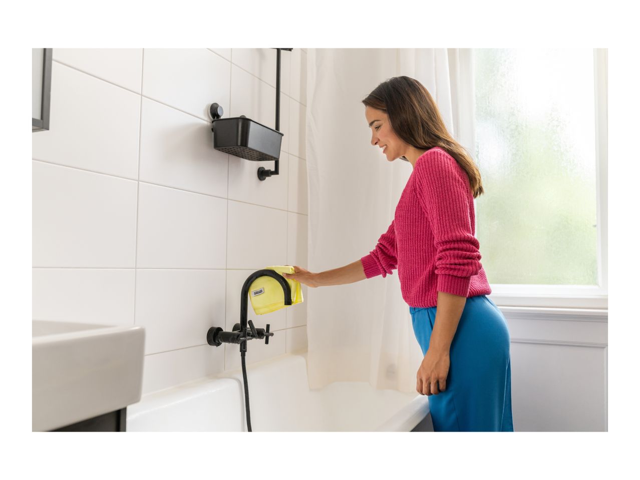 Woman cleaning a black shower faucet with a yellow microfiber cloth in a white tiled bathroom.