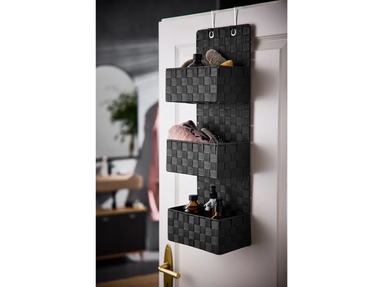 Black hanging organizer with three baskets, holding beauty and personal care products, hanging on a white door.