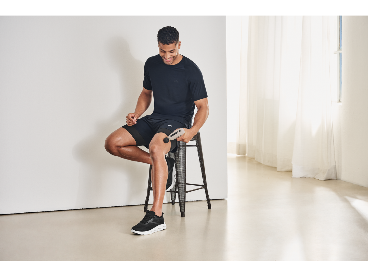 Man using a CRIVIT Mini Massage Gun on his thigh while sitting on a stool.