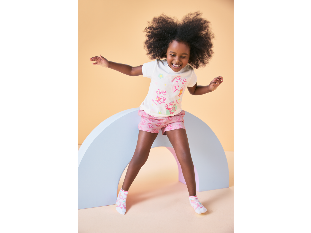 Smiling girl in pink Peppa Pig jersey shorts and matching top, standing over a rainbow prop.