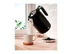 A black electric kettle pouring hot water into a mug on a wooden countertop.