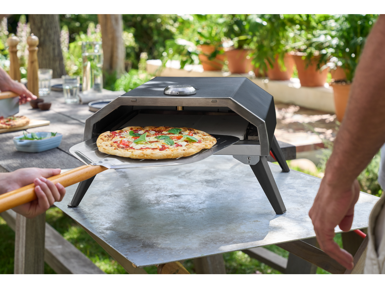 Cuptor de pizza exterior cu o pizza proaspăt coaptă pe o lopată, gata de servit la o petrecere în grădină.