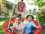 Two happy children in a red swing, with an adult standing behind them in a garden.
