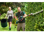 Man trimming a hedge with a long-reach hedge trimmer, woman carrying a garden waste bag.