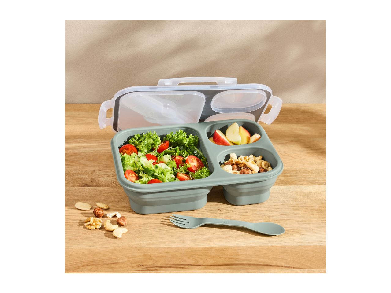 Lunchbox with salad, apple slices, nuts, and a spork on a wooden table.