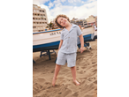 Smiling boy in a light blue short-sleeved shirt and shorts, standing barefoot on a sandy beach.