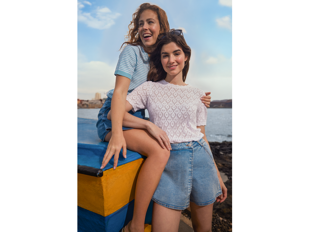 Două femei zâmbitoare poartă tricouri Esmara și pantaloni scurți din denim, lângă mare.
