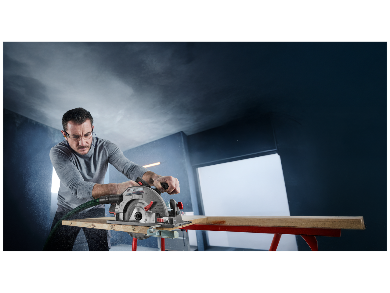 Homme utilisant une scie circulaire pour couper une planche de bois, avec de la poussière en suspension.