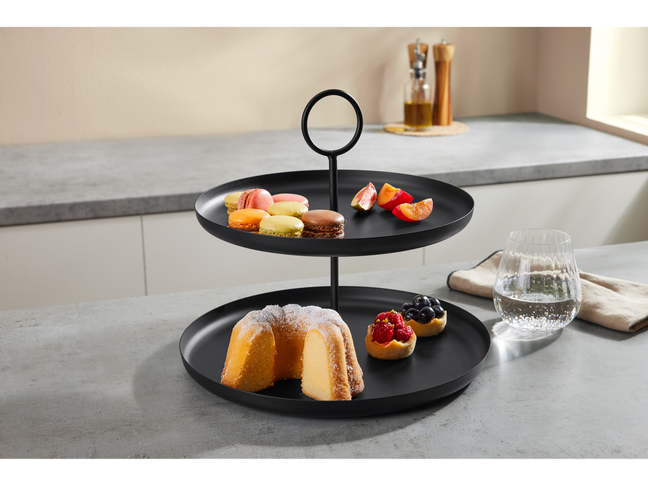 A black two-tiered stand with cakes, macarons, and fruit on a kitchen counter.
