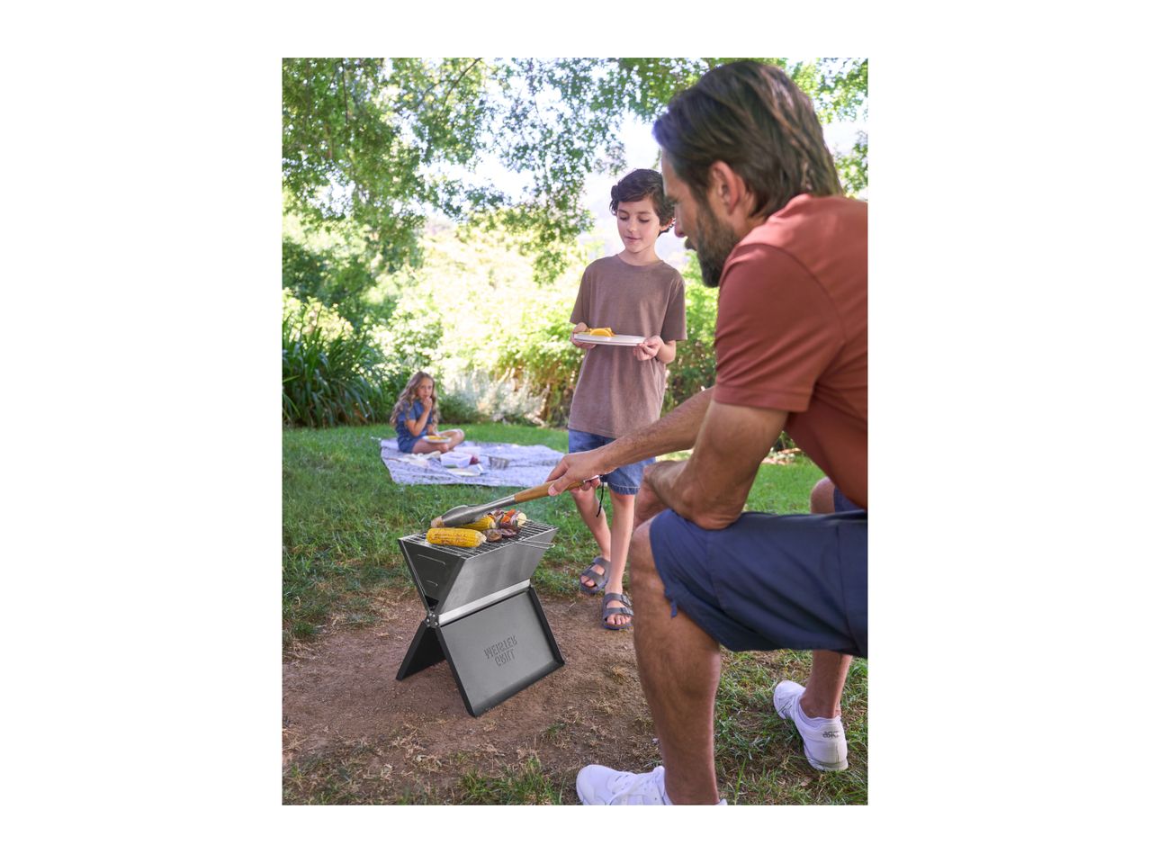 A family barbecuing in a garden, with a portable grill in the foreground.