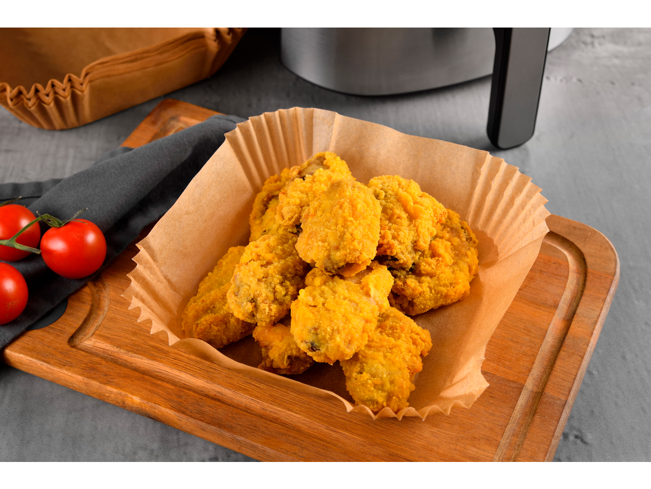 Nuggets de frango em forma de papel sobre uma tábua de madeira com tomates e uma fritadeira de ar.