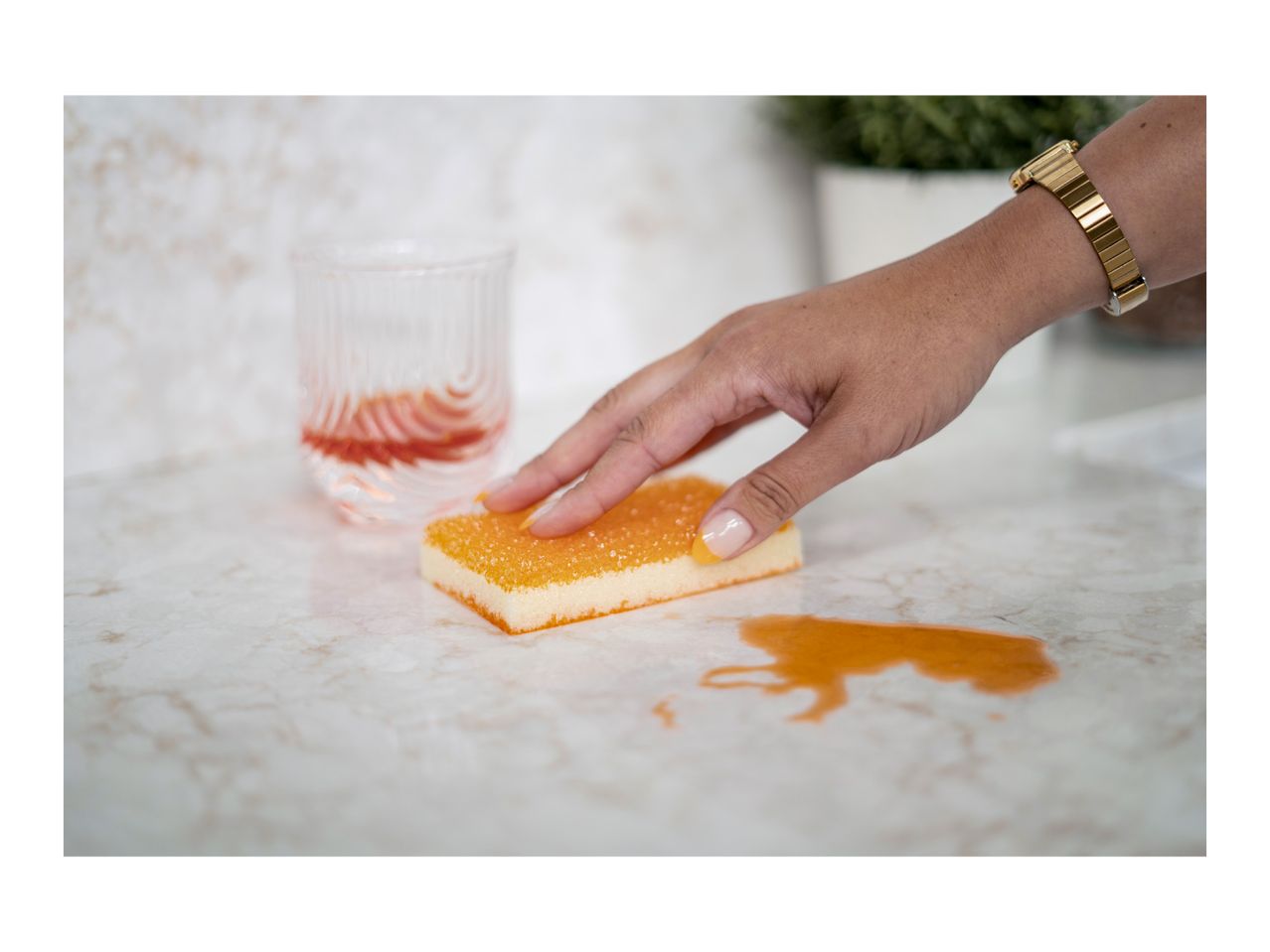 Hand cleaning a spilled drink on a countertop with an orange sponge.