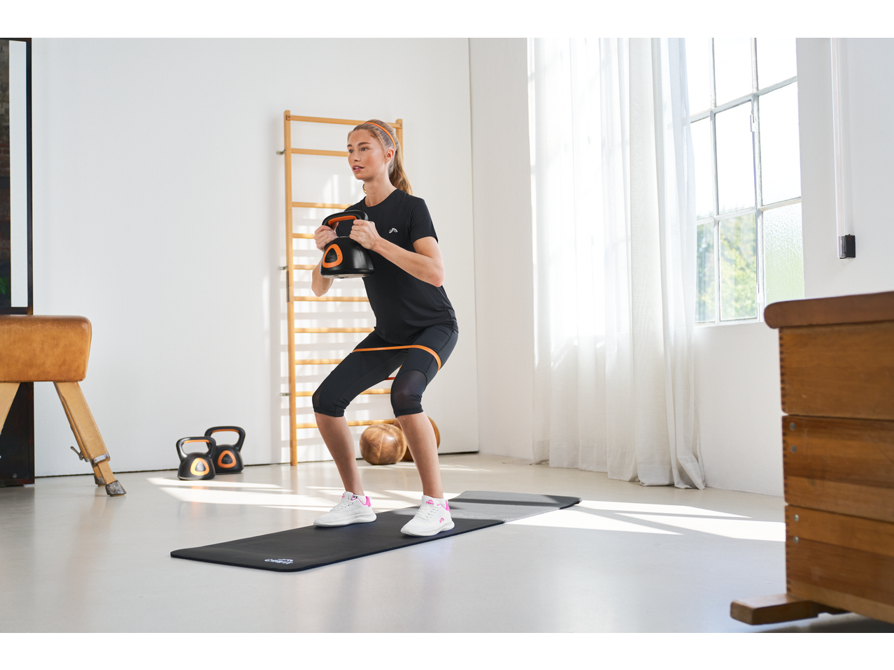 Woman in CRIVIT sportswear doing squats with a kettlebell, resistance band on thighs.