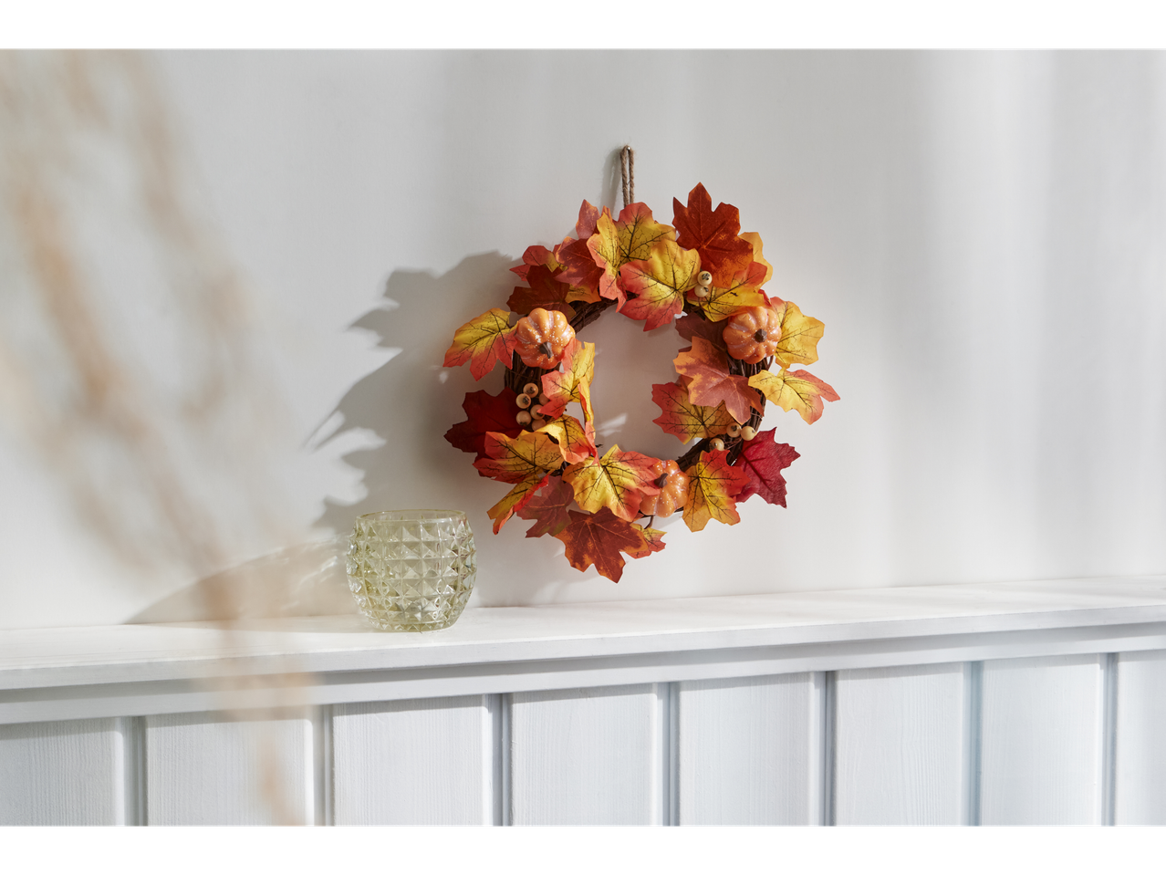 Autumn wreath with pumpkins and leaves, and a glass candle holder on a white shelf.