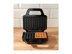 Open waffle maker with one cooked waffle on a wooden surface against a tiled wall.