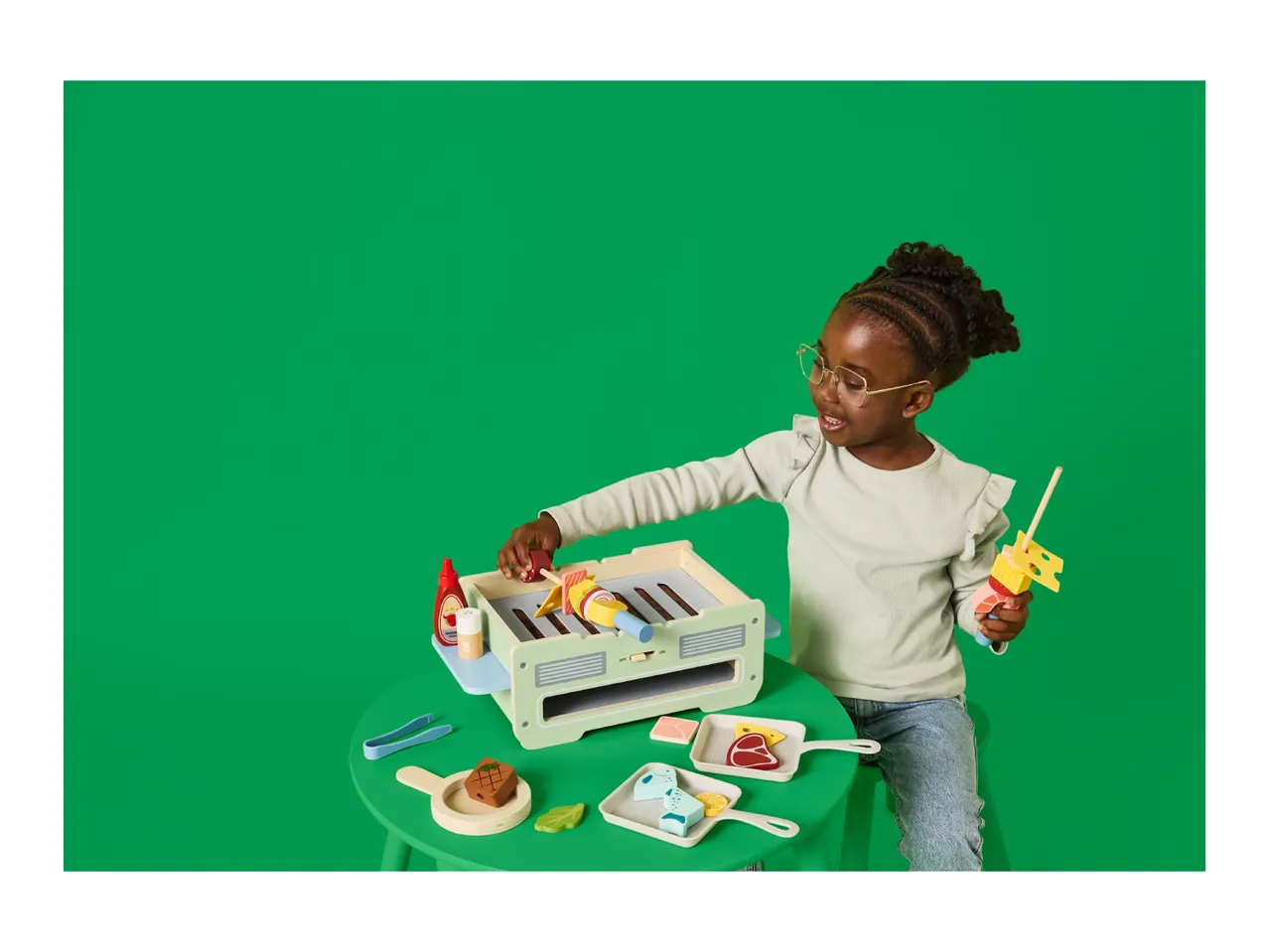 Child playing with a toy barbecue grill and food accessories on a green table.